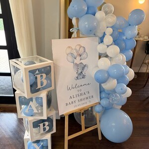 Pode incluir: Uma decora&ccedil;&atilde;o de ch&aacute; de beb&eacute; com uma pilha de quatro caixas brancas transparentes cheias de bal&otilde;es azuis que formam a palavra BABY. Ao lado, um cavalete de madeira sustenta uma placa com uma ilustra&ccedil;&atilde;o de elefante e o texto Welcome TO ALISHA'S BABY SHOWER 29.03.2026, acentuado por um grande cacho de bal&otilde;es azuis e brancos.