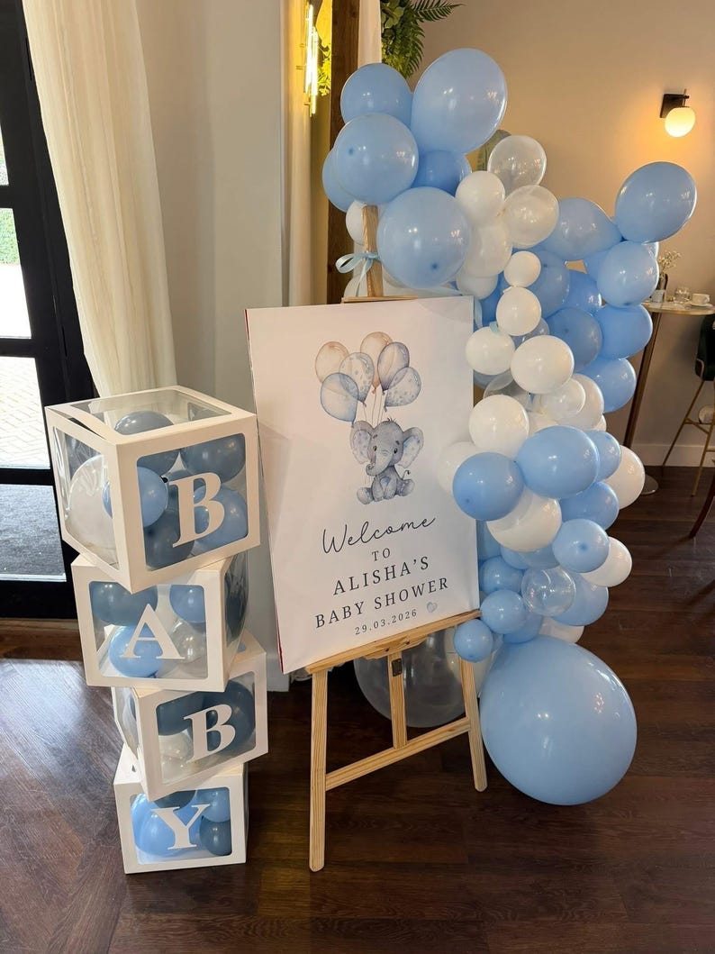 Pode incluir: Uma decora&ccedil;&atilde;o de ch&aacute; de beb&eacute; com uma pilha de quatro caixas brancas transparentes cheias de bal&otilde;es azuis que formam a palavra BABY. Ao lado, um cavalete de madeira sustenta uma placa com uma ilustra&ccedil;&atilde;o de elefante e o texto Welcome TO ALISHA'S BABY SHOWER 29.03.2026, acentuado por um grande cacho de bal&otilde;es azuis e brancos.