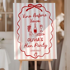May include: A wooden easel displays a sign with a pink and white striped background. The sign features red text and illustrations, including the words "Vino Before Vows" and "Olivia's Hen Party". The date 28.09.2025 is also displayed.