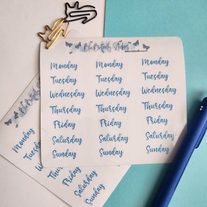 May include: A sheet of clear stickers with the days of the week written in blue cursive lettering. The stickers are arranged in three columns, with each column containing the days of the week from Monday to Sunday.