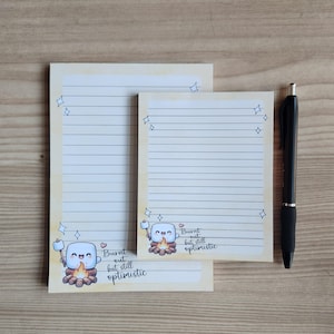 Może przedstawiać: Dwa kremowe, liniowane notatniki z postacią z kreskówki pianki marshmallow i napisem "Burnt out but still optimistic". Notatniki mają jasnożółtą obwódkę i bazgroły gwiazd. Czarny długopis spoczywa na drewnianej powierzchni.