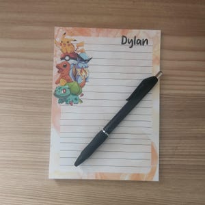 May include: A lined notepad with a watercolor-style orange and peach border. The notepad features a colorful graphic of Pokemon characters and the name "Dylan" in black script. A black pen rests on the notepad.