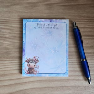 Puede incluir: Bloc de notas con un borde floral azul y el texto «Cosas que olvidaré si no las escribo». Una vaca de dibujos animados con una corona floral está en la esquina. Un bolígrafo azul descansa junto al bloc de notas.