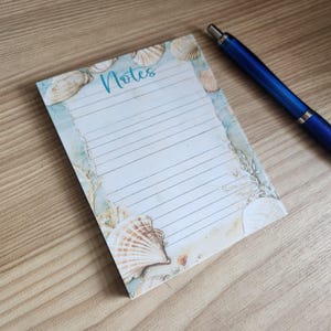 以下が含まれることがあります： 貝殻のデザインとターコイズ色の筆記体で「Notes」という文字が入った長方形のメモ帳。 メモ帳には罫線入りのページがあり、隣の木製の表面には青いペンが置かれています。 メモ帳の縁には貝殻とビーチの風景が描かれています。