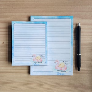 May include: Two lined notepads with a flamingo and duck graphic, and the text "My give a Ducks are on Vacation". The notepads have a blue watercolor border and are next to a black pen.