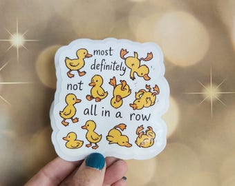 May include: A white sticker with the text "most definitely not all in a row" and illustrations of yellow cartoon ducks in various poses. The sticker is held by a hand with a teal painted fingernail.
