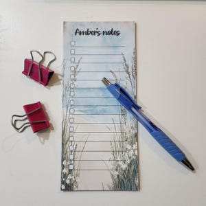 Może przedstawiać: Notatnik z napisem "Amber's notes" i tłem z motywem plaży, niebieski długopis i dwa różowe klipsy. Notatnik ma format listy kontrolnej. Długopis jest niebieski ze srebrną końcówką.