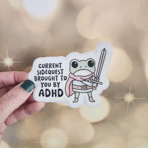 May include: A white sticker with a cartoon frog holding a sword. The frog is wearing a pink scarf and a white belt. The text on the sticker reads "Current Sidequest Brought To You By ADHD".