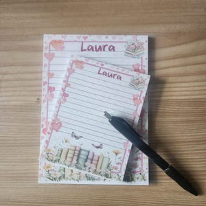May include: Two lined notepads with the name "Laura" printed at the top, decorated with pink hearts and floral illustrations. A black pen rests on the notepad. The notepads feature a floral and book design at the bottom.