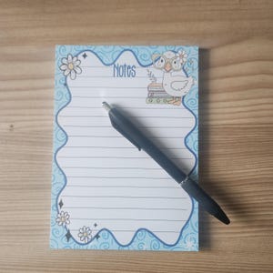Könnte beinhalten: Ein Notizblock mit hellblauem Rand und liniertem Papier. Der Notizblock zeigt das Wort "Notes" und eine Cartoon-Eule mit Brille und Blumenkranz. Ein schwarzer Stift liegt auf dem Notizblock.