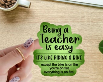 May include: A green sticker with the text "Being a Teacher is easy. It's like riding a bike except the bike is on fire, you're on fire, everything is on fire." The sticker is held by a hand with a gold ring.