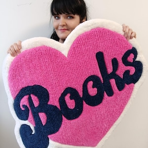 May include: A heart-shaped rug with a pink center and white border. The word "Books" is spelled out in navy blue, cursive lettering across the center. The rug has a soft, textured appearance.