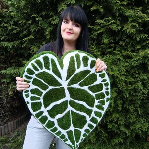 K&ouml;nnte beinhalten: Eine Frau h&auml;lt einen gro&szlig;en, herzf&ouml;rmigen Teppich in Gr&uuml;n und Wei&szlig; mit einem Blattdesign. Der Teppich ist aus einem weichen, flauschigen Material.