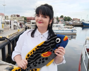 May include: A colorful, handmade fish-shaped rug with a blue head, yellow tail, and a body featuring black, gray, and orange patterns. The rug is held up in front of a harbor with boats and buildings in the background.
