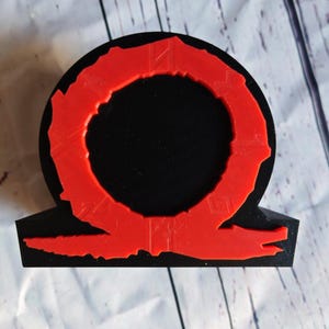 Peut inclure: Une statue en 3D imprim&eacute;e en noir et rouge du logo God of War. Le logo est un cercle avec un anneau rouge autour et une figure rouge en forme de serpent en dessous.