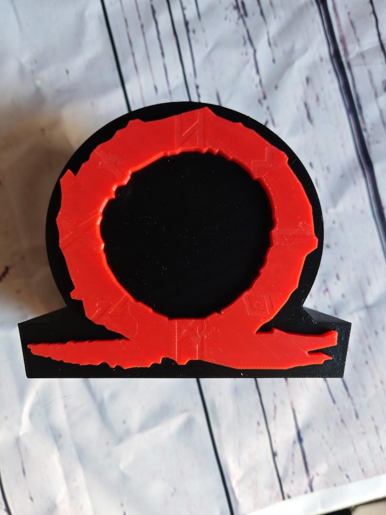 Peut inclure: Une statue en 3D imprim&eacute;e en noir et rouge du logo God of War. Le logo est un cercle avec un anneau rouge autour et une figure rouge en forme de serpent en dessous.