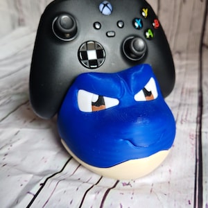 K&ouml;nnte beinhalten: Eine blau-wei&szlig;e Blastoise-Figur mit einem schwarzen Xbox-Controller im Hintergrund. Die Blastoise-Figur hat wei&szlig;e Augen und braune Akzente. Der Controller hat farbige Tasten und ein Schachbrettmuster.