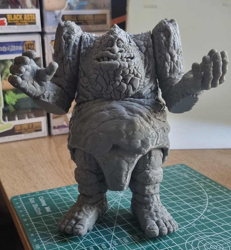 May include: A gray, stone-like figure with a large, toothy mouth and outstretched arms. The figure is standing on a green cutting mat.