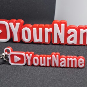 Puede incluir: Llavero impreso en 3D rojo y blanco con un bot&oacute;n de reproducci&oacute;n y el texto "YourName".