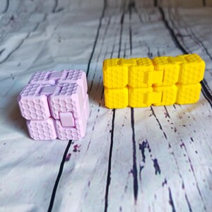 Peut inclure: Deux jouets anti-stress, l'un rose et l'autre jaune. Les deux sont faits d'un mat&eacute;riau en nid d'abeille et ont une forme carr&eacute;e.