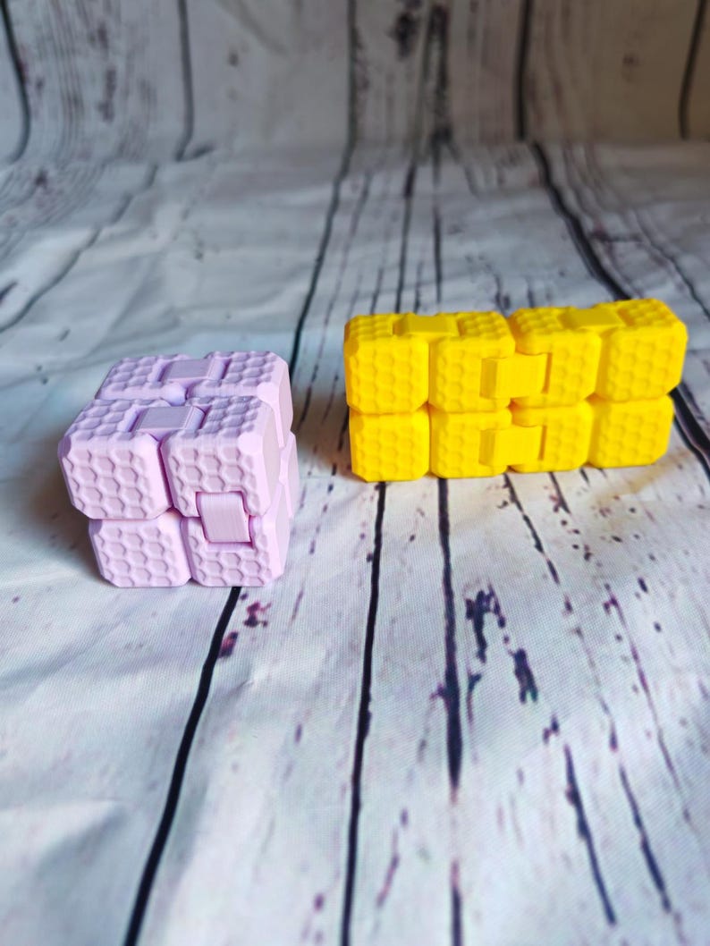 Peut inclure: Deux jouets anti-stress, l'un rose et l'autre jaune. Les deux sont faits d'un mat&eacute;riau en nid d'abeille et ont une forme carr&eacute;e.