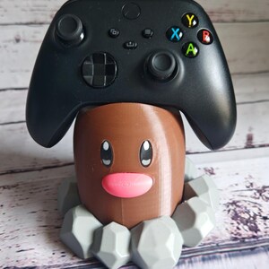 Op de afbeelding: Een 3D-geprinte bruine en grijze Pokemon Diglett controllerstandaard. De standaard heeft een roze neus en zwarte ogen. De standaard houdt een zwarte Xbox-controller.