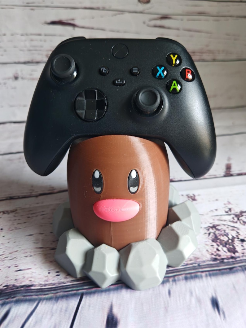 Op de afbeelding: Een 3D-geprinte bruine en grijze Pokemon Diglett controllerstandaard. De standaard heeft een roze neus en zwarte ogen. De standaard houdt een zwarte Xbox-controller.