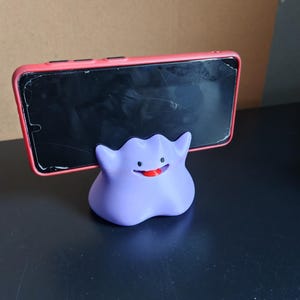 以下が含まれることがあります： ポケモンのキャラクターの形をした、紫と赤の3Dプリントされた携帯電話スタンド。スタンドは、ひび割れた画面を持つ黒いスマートフォンを保持しています。