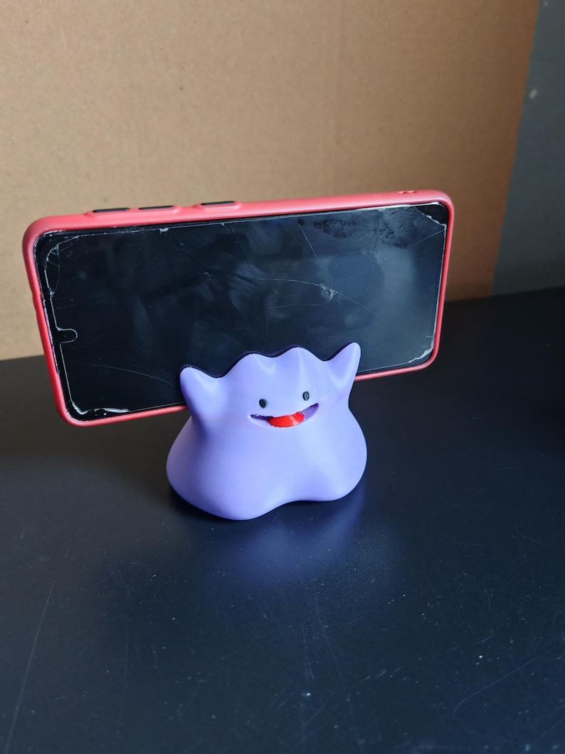 以下が含まれることがあります： ポケモンのキャラクターの形をした、紫と赤の3Dプリントされた携帯電話スタンド。スタンドは、ひび割れた画面を持つ黒いスマートフォンを保持しています。
