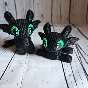 May include: Two black crocheted plush toys shaped like dragons with green eyes. The dragons are sitting on a white wooden surface.