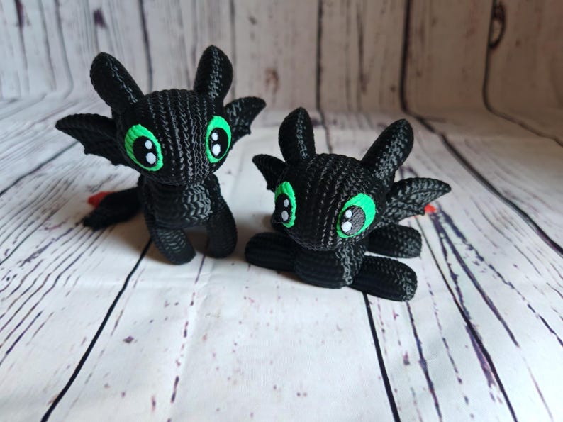 May include: Two black crocheted plush toys shaped like dragons with green eyes. The dragons are sitting on a white wooden surface.