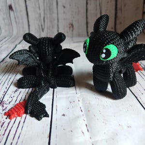 May include: Two black crocheted plush toys shaped like dragons. One dragon has green eyes and is standing. The other dragon is lying down with its wings spread out. Both dragons have red tails.