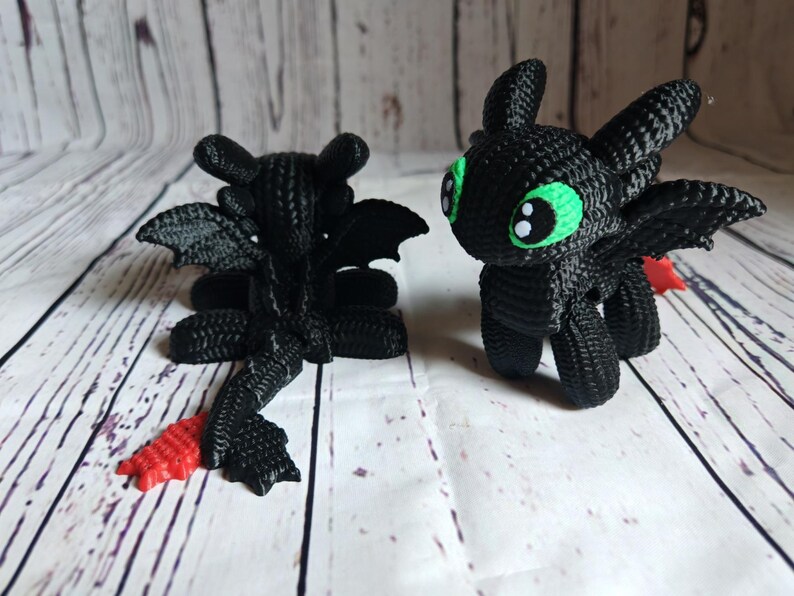 May include: Two black crocheted plush toys shaped like dragons. One dragon has green eyes and is standing. The other dragon is lying down with its wings spread out. Both dragons have red tails.