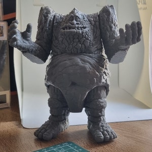 May include: A gray, stone-like figure with a textured surface and a wide, open mouth. The figure has a large, round belly and short, stubby legs. The figure is standing on a wooden surface.