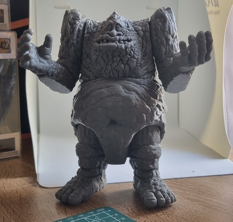 May include: A gray, stone-like figure with a textured surface and a wide, open mouth. The figure has a large, round belly and short, stubby legs. The figure is standing on a wooden surface.