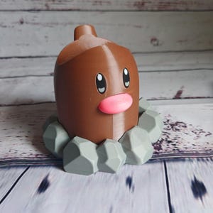 Op de afbeelding: Een bruine en grijze Pokemon-figuur van een Diglett, een grond-type Pokemon. De figuur is gemaakt van plastic en heeft een gedetailleerd ontwerp. De Diglett staat op een basis van grijze stenen.