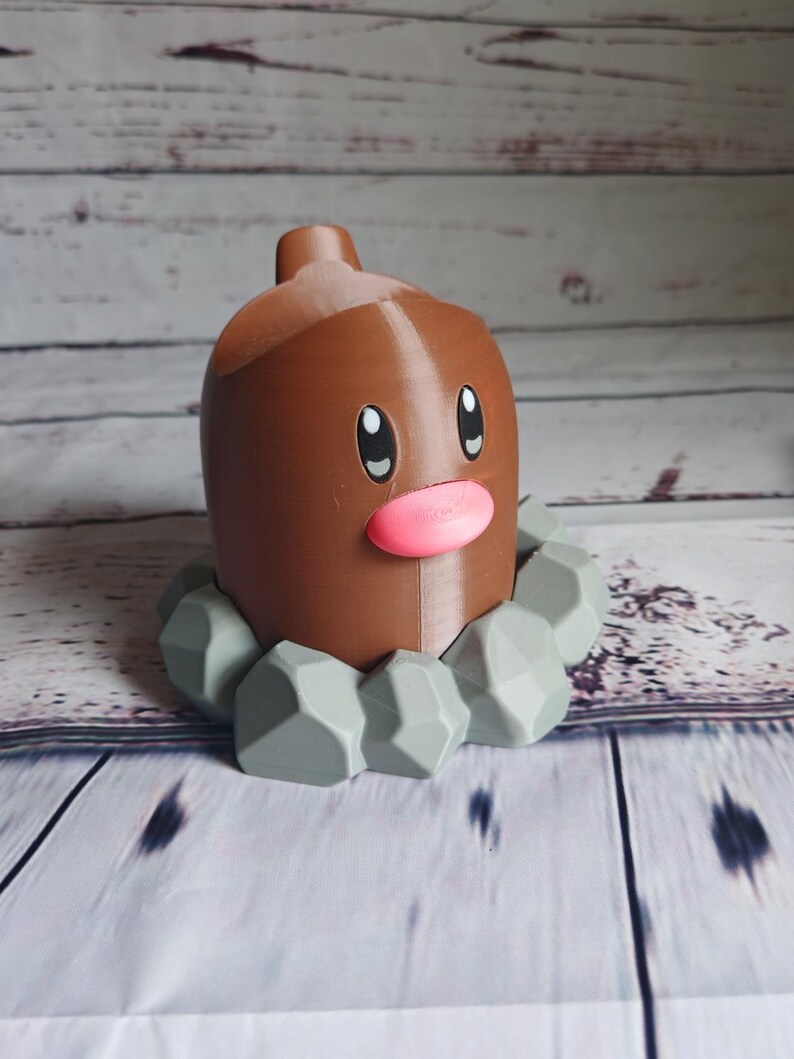 Op de afbeelding: Een bruine en grijze Pokemon-figuur van een Diglett, een grond-type Pokemon. De figuur is gemaakt van plastic en heeft een gedetailleerd ontwerp. De Diglett staat op een basis van grijze stenen.