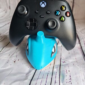 Op de afbeelding: Zwarte Xbox-controller op een lichtblauwe Lapras-standaard. De controller heeft gekleurde knoppen en twee joysticks. De standaard is een gestileerd Pok&eacute;mon-personage.