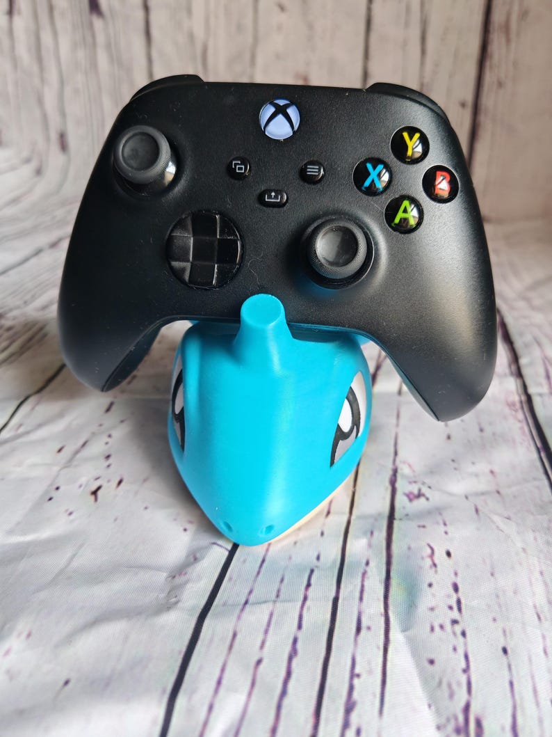 Op de afbeelding: Zwarte Xbox-controller op een lichtblauwe Lapras-standaard. De controller heeft gekleurde knoppen en twee joysticks. De standaard is een gestileerd Pok&eacute;mon-personage.
