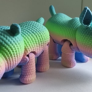 May include: Two colourful, crocheted animal toys. A pink, blue and green hippopotamus and a pink, blue and green rhinoceros.