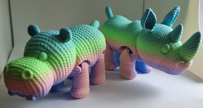 May include: Two colourful, crocheted animal toys. A pink, blue and green hippopotamus and a pink, blue and green rhinoceros.