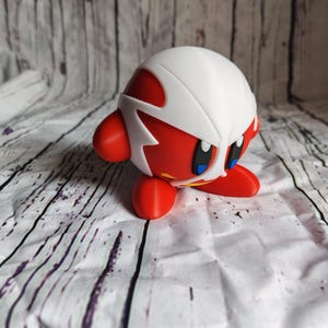 Pode incluir: Estatueta vermelha e branca de um personagem com grandes olhos azuis e uma m&aacute;scara branca. A figura tem bra&ccedil;os e p&eacute;s vermelhos e est&aacute; em posi&ccedil;&atilde;o de c&oacute;coras. O fundo &eacute; uma superf&iacute;cie de madeira branca e cinza.