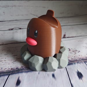 Op de afbeelding: Een 3D-geprint Pokemon-figuur van Diglett, een grond-type Pokemon, met een klein, rond lichaam en een puntige kop. Het figuur staat op een basis van grijze stenen.