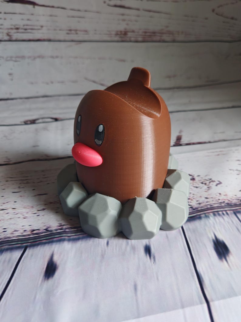 Op de afbeelding: Een 3D-geprint Pokemon-figuur van Diglett, een grond-type Pokemon, met een klein, rond lichaam en een puntige kop. Het figuur staat op een basis van grijze stenen.