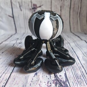 May include: A black and white 3D printed toy with a spider-like design. The toy has a round head with a white Venom symbol and six black legs with wheels.
