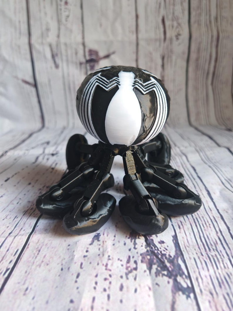 May include: A black and white 3D printed toy with a spider-like design. The toy has a round head with a white Venom symbol and six black legs with wheels.