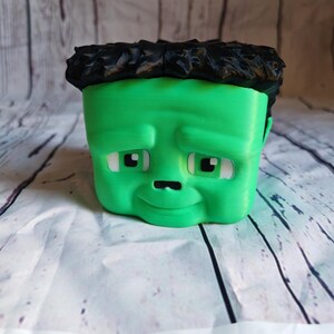 K&ouml;nnte beinhalten: Ein gr&uuml;ner, 3D-gedruckter Pflanztopf in Form von Frankensteins Monster. Der Topf hat ein quadratisches Gesicht, schwarzes, strukturiertes Haar, wei&szlig;e Augen und ein kleines, gebogenes L&auml;cheln. Der Hintergrund ist eine wei&szlig;-graue, holz&auml;hnliche Oberfl&auml;che.