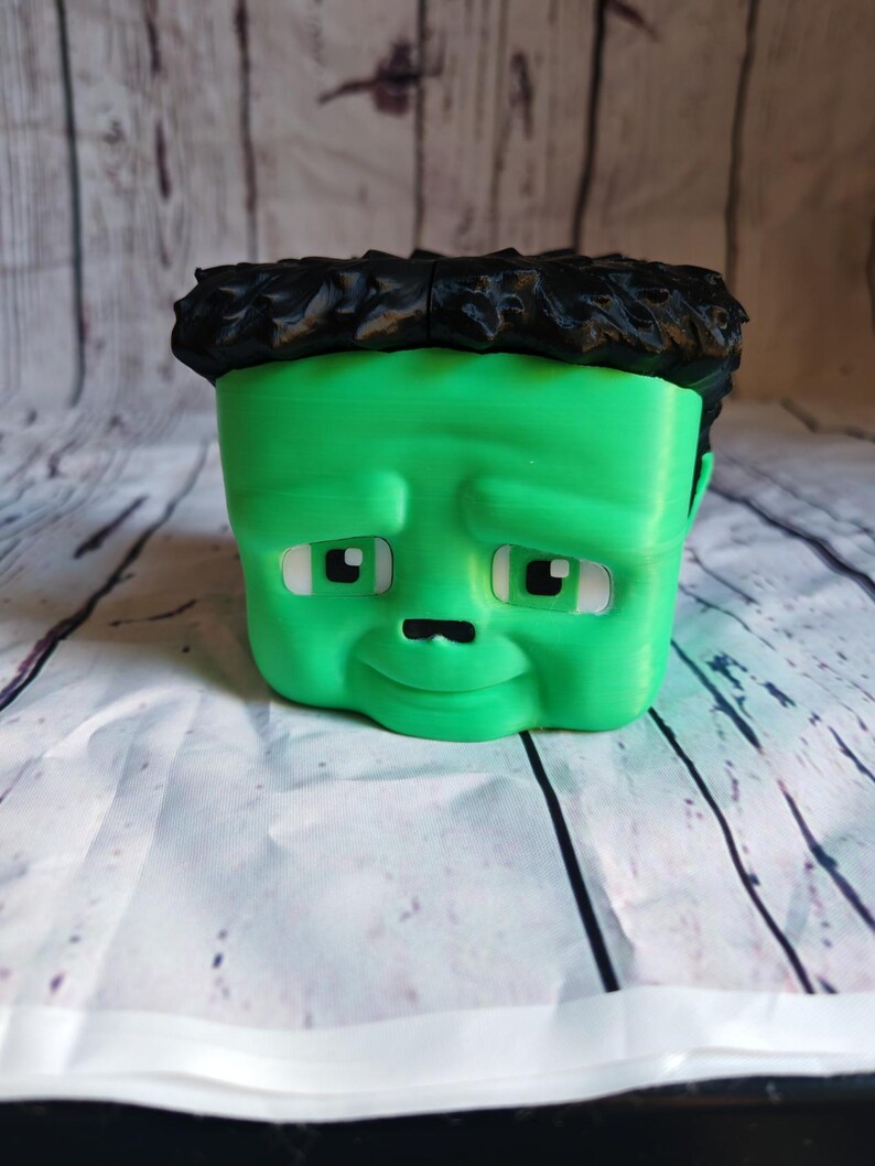 K&ouml;nnte beinhalten: Ein gr&uuml;ner, 3D-gedruckter Pflanztopf in Form von Frankensteins Monster. Der Topf hat ein quadratisches Gesicht, schwarzes, strukturiertes Haar, wei&szlig;e Augen und ein kleines, gebogenes L&auml;cheln. Der Hintergrund ist eine wei&szlig;-graue, holz&auml;hnliche Oberfl&auml;che.