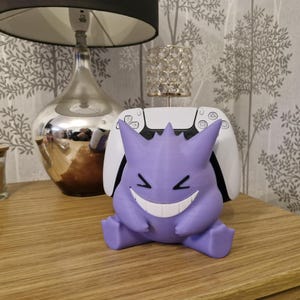 May include: A purple and white 3D printed Gengar figurine with a wide grin, sitting on a white gaming console.