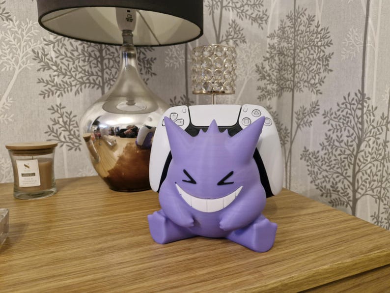 May include: A purple and white 3D printed Gengar figurine with a wide grin, sitting on a white gaming console.